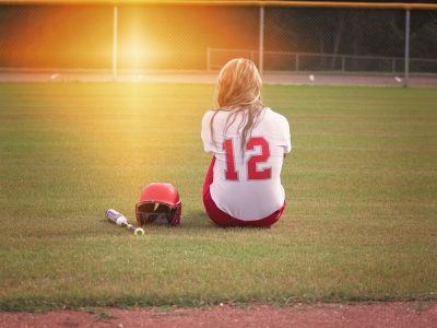 softball player facing away, sitting on the ground, with a bat and helmet beside her